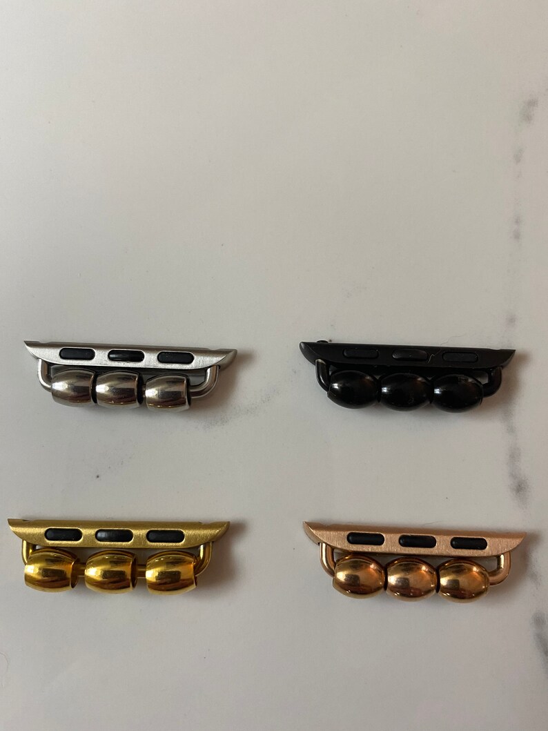 K&ouml;nnte beinhalten: Vier Apple Watch Band-Adapter in verschiedenen Ausf&uuml;hrungen: Silber, Schwarz, Gold und Ros&eacute;gold. Jeder Adapter hat drei runde Perlen und einen rechteckigen Einsatz mit vier schwarzen rechteckigen Ausschnitten.