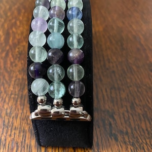 K&ouml;nnte beinhalten: Ein dreireihiges Armband aus Fluoritperlen in Gr&uuml;n-, Blau- und Violettt&ouml;nen. Das Armband ist f&uuml;r eine Apple Watch konzipiert und hat einen silbernen Metallverschluss.