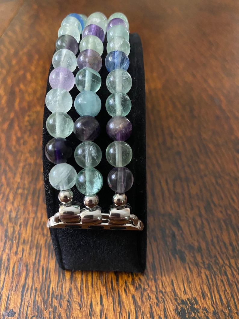 K&ouml;nnte beinhalten: Ein dreireihiges Armband aus Fluoritperlen in Gr&uuml;n-, Blau- und Violettt&ouml;nen. Das Armband ist f&uuml;r eine Apple Watch konzipiert und hat einen silbernen Metallverschluss.