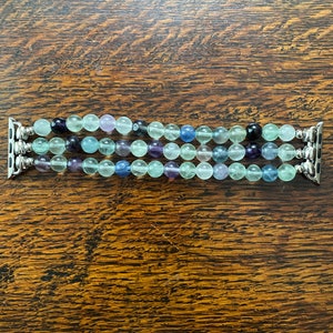 K&ouml;nnte beinhalten: Ein mehrreihiges Apple Watch Armband mit gr&uuml;nen, blauen und violetten Perlen. Das Armband besteht aus nat&uuml;rlichen Steinen und hat einen silbernen Metallverschluss.