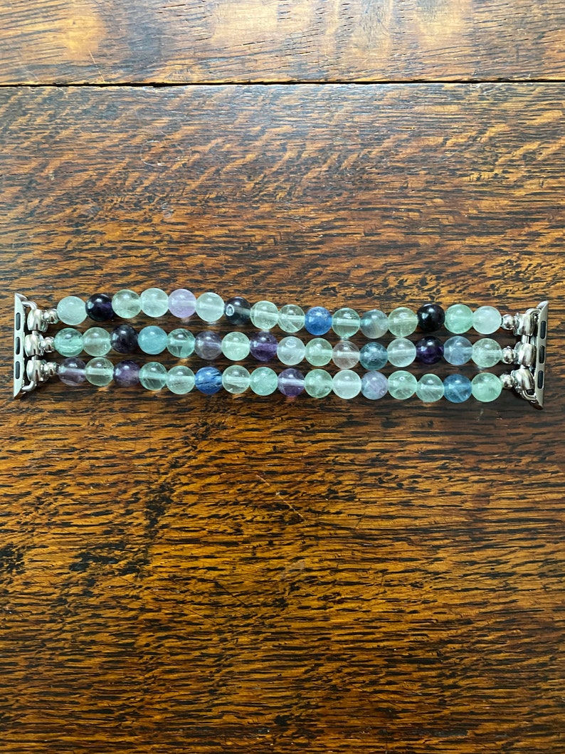 K&ouml;nnte beinhalten: Ein mehrreihiges Apple Watch Armband mit gr&uuml;nen, blauen und violetten Perlen. Das Armband besteht aus nat&uuml;rlichen Steinen und hat einen silbernen Metallverschluss.