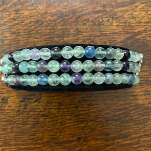 K&ouml;nnte beinhalten: Ein mehrreihiges Armband aus gr&uuml;nen, blauen und violetten Edelsteinperlen. Die Perlen sind rund und haben eine glatte, polierte Oberfl&auml;che. Das Armband liegt auf einem schwarzen Samt-Hintergrund.
