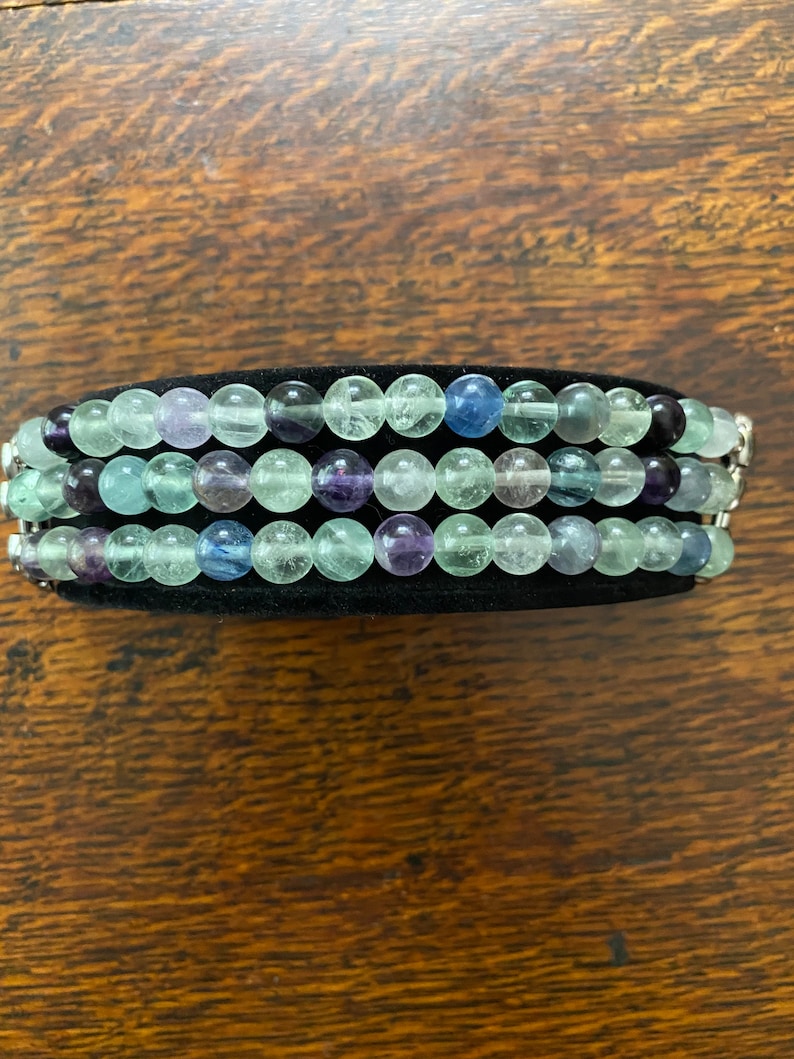 K&ouml;nnte beinhalten: Ein mehrreihiges Armband aus gr&uuml;nen, blauen und violetten Edelsteinperlen. Die Perlen sind rund und haben eine glatte, polierte Oberfl&auml;che. Das Armband liegt auf einem schwarzen Samt-Hintergrund.