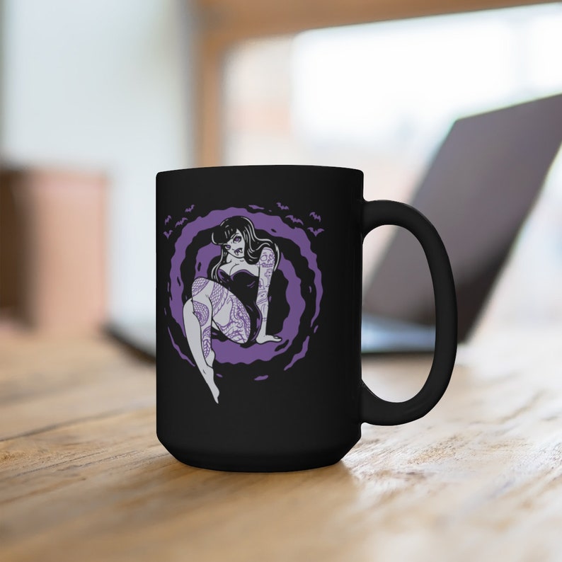 K&ouml;nnte beinhalten: Schwarze Keramiktasse mit einer lila und wei&szlig;en Illustration einer t&auml;towierten Frau, die in einer Wolke aus Flederm&auml;usen sitzt.