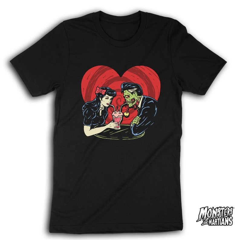 May include: Black t-shirt with a graphic of a zombie and a woman sharing a milkshake in a heart-shaped frame. The text "Monsters and Martians" is in the bottom right corner.