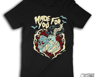 Peut inclure: Un t-shirt noir à manches courtes avec une impression graphique représentant un personnage à la peau bleue aux cheveux foncés tenant un crâne cousu. Le texte "MADE FOR YOU" est inscrit en crème, entouré de toiles d'araignée rouges et de chauves-souris bleues.