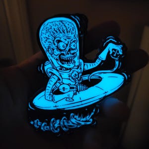 May include: A glow-in-the-dark enamel pin featuring a Martian character with an exposed brain, wearing a space suit, and holding a ray gun while emerging from a flying saucer. The design is rendered in bright cyan blue against a black background.