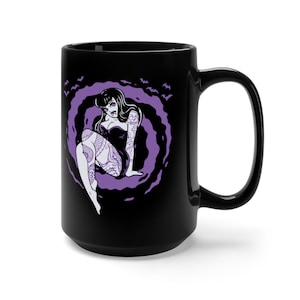 K&ouml;nnte beinhalten: Schwarze Keramiktasse mit einer lilafarbenen Illustration einer t&auml;towierten Frau, die in einer wirbelnden Wolke sitzt. Flederm&auml;use fliegen um die Wolke herum.