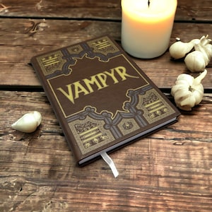 May include: A brown hardback book with the word VAMPYR printed in large gold letters on the front cover. The cover features intricate gold and blue decorative border patterns and a white ribbon bookmark extending from the bottom edge.