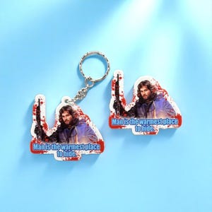 May include: Two acrylic keyrings featuring a man holding a rifle, with the text "Man is the warmest place to hide." The keyrings have a red and white splatter design and are attached to a silver keyring. The background is light blue.