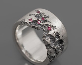 Può includere: Un anello d'argento con una superficie strutturata e piccole pietre preziose rosse. L'anello presenta un design organico e irregolare, con una texture ruvida e irregolare sulla fascia esterna. Le pietre rosse sono raggruppate in un motivo floreale.