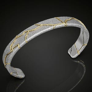 May include: Silver-tone cuff bracelet with gold-colored accents. The bracelet has a textured surface with gold-colored lines running across it. The ends of the bracelet are smooth silver. The jewelry is displayed on a black surface.