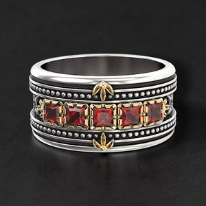 May include: A silver ring with a black band, adorned with five square, red gemstones. The ring features gold-colored accents and small, silver beads, creating a detailed and ornate design.