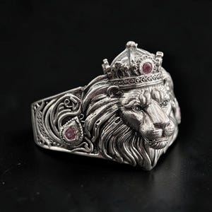Könnte beinhalten: Ein silberfarbener Ring mit einem detaillierten Löwenkopf, der eine Krone trägt. Der Löwe ist mit kleinen, runden, rosafarbenen Steinen besetzt, die in die Krone und die Seite des Rings eingelassen sind. Das Metall hat ein oxidiertes Finish.