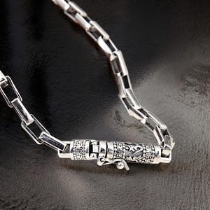 May include: Close-up of a silver chain necklace with a decorative clasp. The chain links are rectangular and connected. The clasp is cylindrical with engraved patterns. The necklace is displayed on a dark surface.