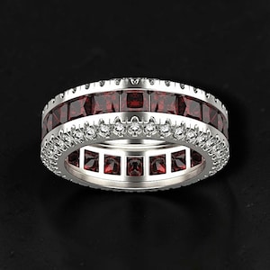 May include: A silver eternity band featuring a central row of square-cut deep red gemstones flanked by two rows of small, round, clear white stones. The metal band has a polished finish and a consistent width throughout its circular design.