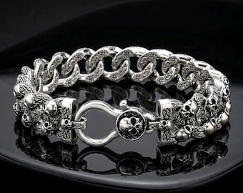 May include: A silver-toned metal chain bracelet featuring a repeating pattern of small skulls integrated into the links. The bracelet includes a circular lobster claw clasp mechanism that is also adorned with a detailed skull design.