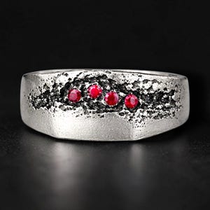 May include: A silver ring with a textured band and four small, bright red gemstones. The ring has a rugged, almost stone-like appearance, with a dark, textured groove running across the top where the gems are set.