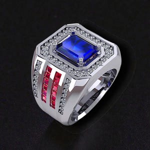 May include: A silver ring with a large, rectangular blue gemstone at the center, surrounded by smaller clear stones. The ring features three vertical red gemstone accents on each side and is set against a black background.