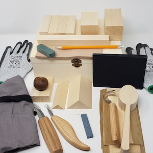 May include: A wood carving kit containing a wooden box, carving tools, wood blanks, a sharpening stone, gloves, and a pencil. The box is made of light wood and has a brass latch. The carving tools are made of wood and metal. The wood blanks are of various shapes and sizes. The sharpening stone is grey and rectangular. The gloves are grey and have green accents. The pencil is yellow. The kit is ideal for beginners or experienced wood carvers.