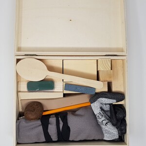 May include: A wooden box with a lid open, revealing a variety of items inside. The items include a wooden spoon, a green sponge, a grey sanding block, a brown leather strap, a pencil, a brown ball, and a grey work glove.