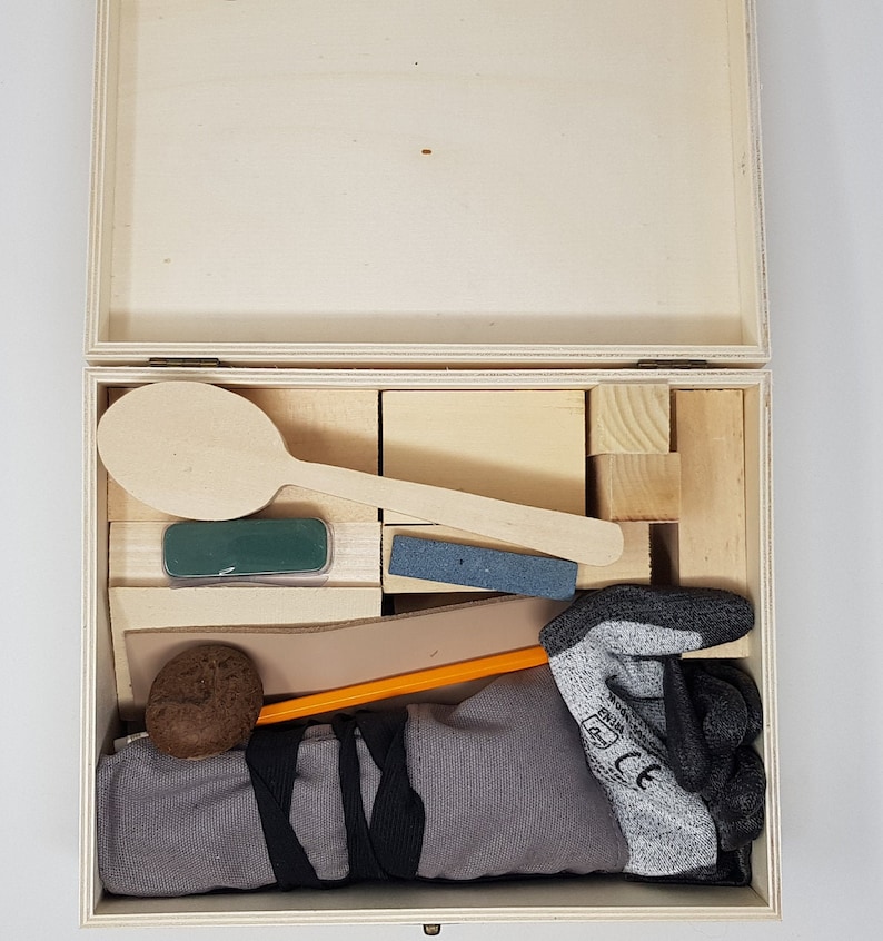 May include: A wooden box with a lid open, revealing a variety of items inside. The items include a wooden spoon, a green sponge, a grey sanding block, a brown leather strap, a pencil, a brown ball, and a grey work glove.