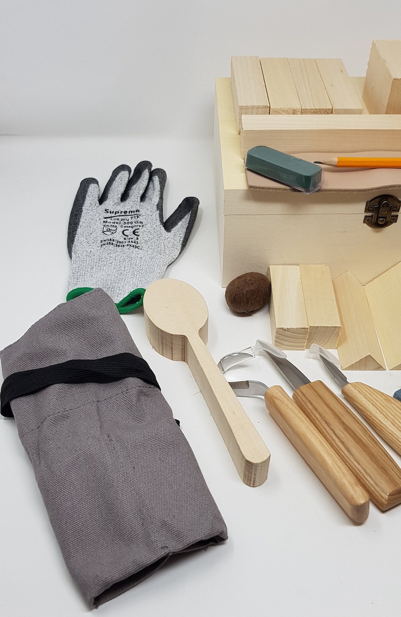 May include: A wood carving kit containing a wooden spoon, carving tools, and a wooden box with wooden blocks. The kit also includes a pair of grey work gloves and a grey canvas roll.