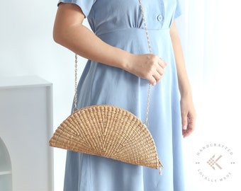 Könnte beinhalten: Ein hellblaues Kleid mit einer goldenen Kettenriementasche aus geflochtenem Stroh. Die Tasche hat eine Halbkreisform und ist mit einem goldenen Verschluss verziert. Der Text "HANDCRAFTED LOCALLY MADE" ist in der unteren rechten Ecke des Bildes sichtbar.