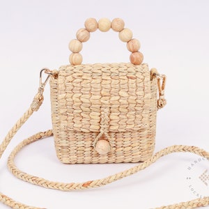 May include: A small, woven straw bag with a wooden bead handle and a long, woven strap. The bag has a flap closure and a small wooden bead accent.