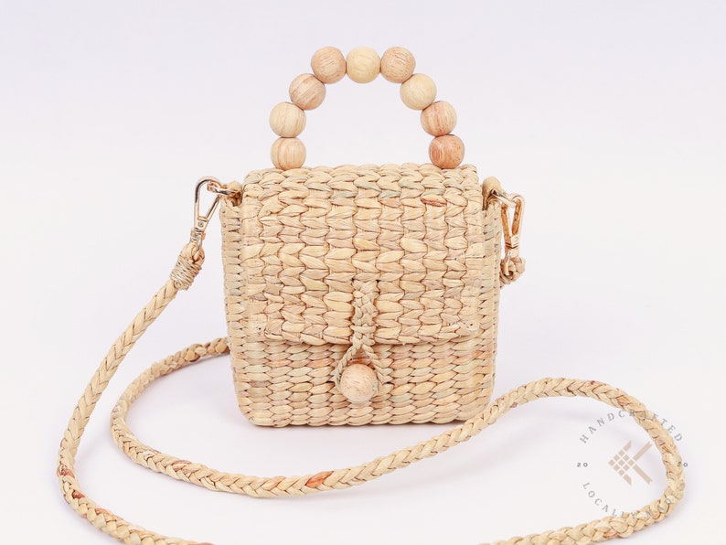 May include: A small, woven straw bag with a wooden bead handle and a long, woven strap. The bag has a flap closure and a small wooden bead accent.