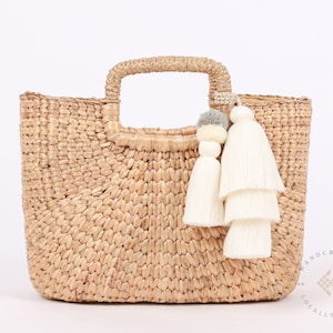 May include: A natural woven straw tote bag with a brown handle and white tassel accents. The bag has a "HANDCRAFTED LOCALLY MADE" label on the bottom.