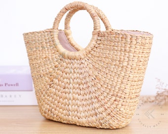 May include: A natural woven straw tote bag with a large, rounded, open top and a single, large, circular handle. The bag is made of natural straw and has a light brown colour. The bag is sitting on a wooden surface. The text "HANDCRAFTED LOCALLY MADE" is visible on the bottom right corner of the image.