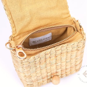 May include: A small, woven straw bag with a zipper closure and a gold metal zipper pull. The bag has a brown fabric lining and a wooden button closure. The bag is handcrafted and locally made.