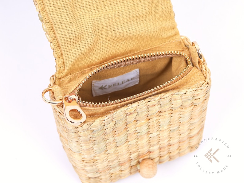 May include: A small, woven straw bag with a zipper closure and a gold metal zipper pull. The bag has a brown fabric lining and a wooden button closure. The bag is handcrafted and locally made.