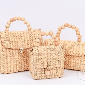 May include: Three woven straw handbags with wooden bead handles. The largest bag has a flap closure with a gold clasp. The smallest bag has a single wooden bead on the flap.