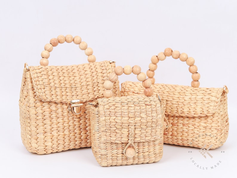 May include: Three woven straw handbags with wooden bead handles. The largest bag has a flap closure with a gold clasp. The smallest bag has a single wooden bead on the flap.