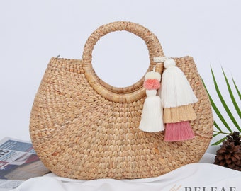Könnte beinhalten: Eine gewebte Strohhandtasche mit rundem Griff und dekorativem Quastenanhänger. Die Tasche hat eine hellbraune Farbe und eine abgerundete Form. Der Quastenanhänger ist weiß, rosa und beige. Ein stilvolles Accessoire.