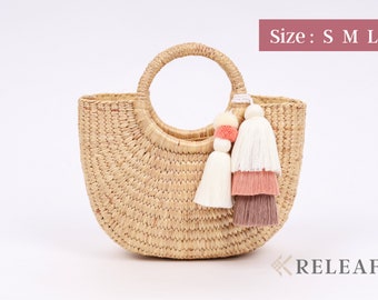 May include: A half-moon shaped straw tote bag with a round handle and a tassel detail. The tassel is made of white, pink, and brown yarn. The bag is available in small, medium, and large sizes. Size: S M L