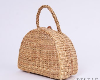 Könnte beinhalten: Eine hellbraune geflochtene Strohtasche mit abgerundetem, kuppelförmigem Oberteil und einem einzigen geflochtenen Henkel. Die Tasche hat einen Verschluss aus Holz.