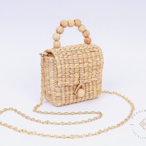 May include: A small, woven straw bag with a wooden bead handle and a gold chain strap. The bag has a flap closure and a small wooden button. The bag is handcrafted and locally made.