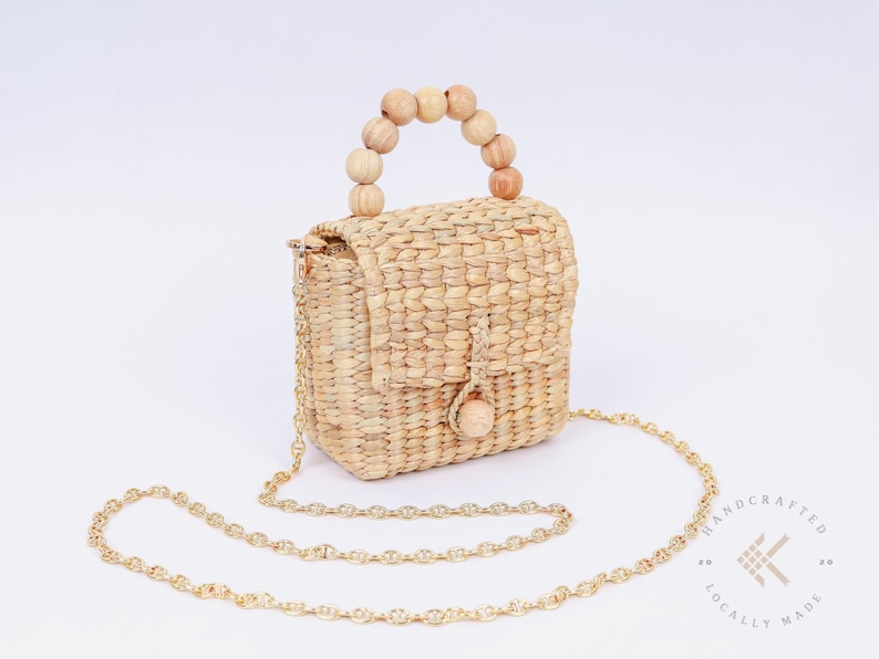 May include: A small, woven straw bag with a wooden bead handle and a gold chain strap. The bag has a flap closure and a small wooden button. The bag is handcrafted and locally made.