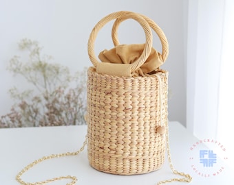 May include: A woven straw bucket bag with a brown leather strap and a brown fabric lining. The bag has a gold chain strap and is handcrafted and locally made.