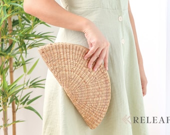 May include: A woman is holding a half-moon shaped woven clutch bag made from natural materials. The bag is light brown and has a woven pattern. The woman is wearing a light green dress.