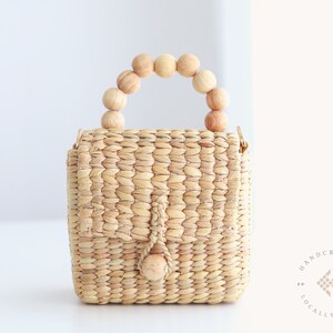May include: A small, woven straw handbag with a wooden bead handle. The bag has a flap closure and a small wooden bead accent. The bag is handcrafted and locally made.