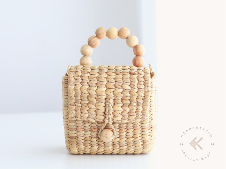 May include: A small, woven straw handbag with a wooden bead handle. The bag has a flap closure and a small wooden bead accent. The bag is handcrafted and locally made.