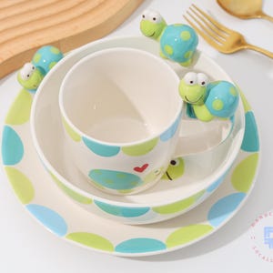 Könnte beinhalten: Ein weißes Keramik-Kindergeschirr-Set mit Schildkröten-Motiv. Das Set enthält einen Teller, eine Schüssel und eine Tasse, alle mit blauen und grünen Tupfen und Schildkröten-Akzenten verziert. Ein Holzschneidebrett und goldenes Besteck befinden sich im Hintergrund.