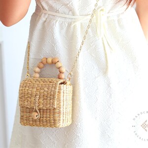 May include: A small, woven straw bag with a wooden handle. The bag is hanging from a gold chain strap and is being worn by a person wearing a white dress. The bag has a flap closure and a small wooden knob.