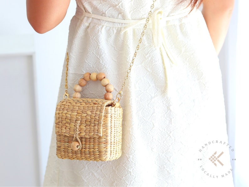 May include: A small, woven straw bag with a wooden handle. The bag is hanging from a gold chain strap and is being worn by a person wearing a white dress. The bag has a flap closure and a small wooden knob.