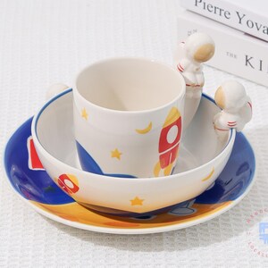 May include: A white ceramic children's dish set with a space theme. The set includes a cup, bowl, and plate, decorated with rockets, stars, and moons. The cup has astronaut figures as handles. The set is designed for children.