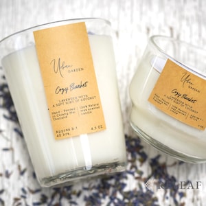 Op de afbeelding: Twee glazen kaarsenpotten met een beige label waarop staat "Urban Garden Cozy Blanket Lavender with a soft hint of coconut" en "Hand-Poured in Chiang Mai Thailand 100% Natural wax scented candle Approx b.t 40 hrs. 4.5 OZ". De kaarsenpotten staan op een wit houten oppervlak met gedroogde lavendelbloemen.